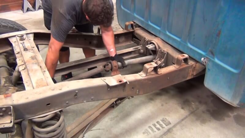 A mechanic pulling the driveshaft out of a classic Chevy truck through the area underneath the missing truck bed