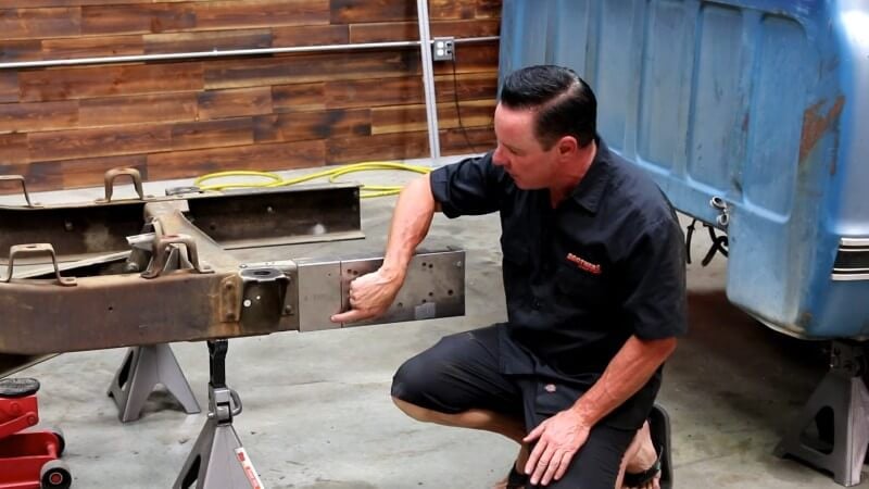 A mechanic kneeling down next to the rear frame section and pointing at the holes that need to be drilled on the template