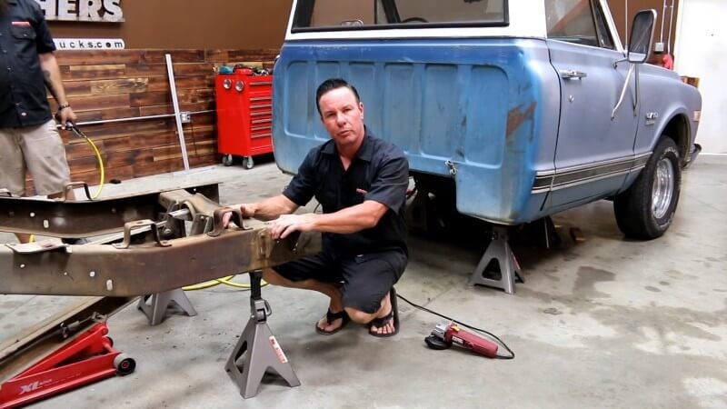 A mechanic kneeling in front of the classic Chevy truck and resting his hand on the forward bed mount that needs to be removed