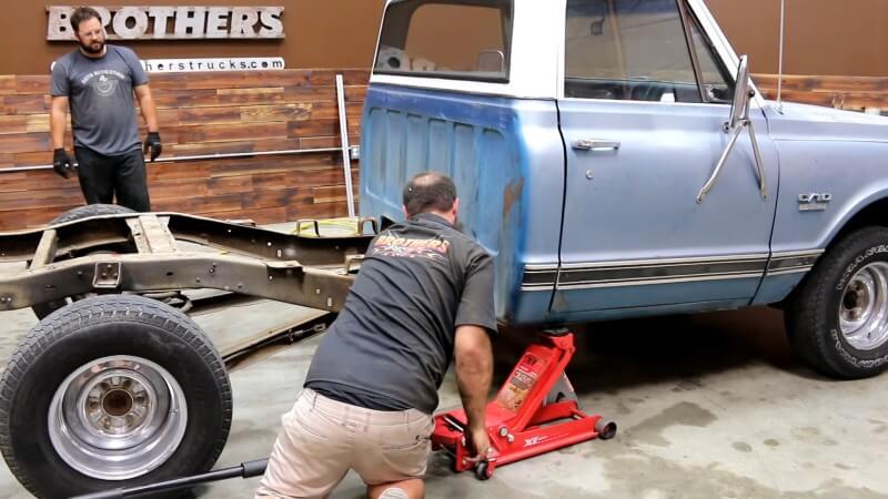 Two mechanics using a red hydraulic floor jack to lift up the rear of the truck's cab