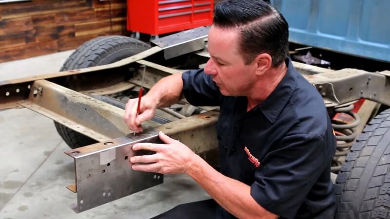 A mechanic using kneeling in front of the rear frame section and marking where the rear bed mount hole should be on the modified template