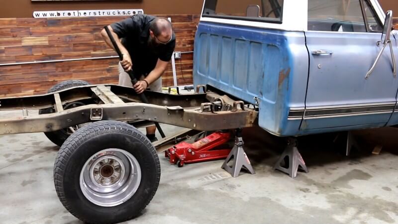 A mechanic positioning a red hydraulic floor jack underneath the now cut section of the rear crossmember