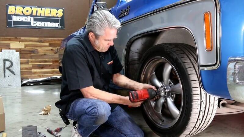A mechanic kneeling next to a classic Chevy truck while using an impact wrench to remove the lug nuts on the front passenger side wheel