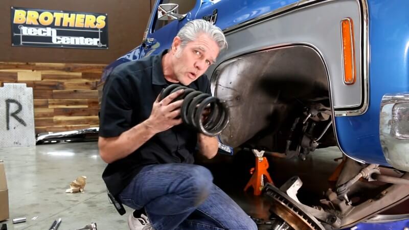 A mechanic kneeling next to a classic Chevy truck while holding up the old coil spring to the camera