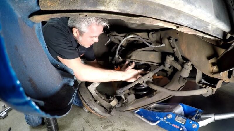 A mechanic kneeling under a classic Chevy truck while moving the new coil spring into position on the lower control arm