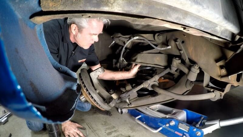 A mechanic kneeling under a classic Chevy truck while holding the new coil spring into place as another mechanic off screen raises the lower control arm with a jack
