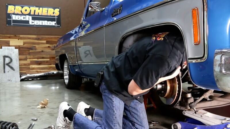 A mechanic kneeling under a classic Chevy truck while leaning over the brake rotor and reconnecting the upper ball joint