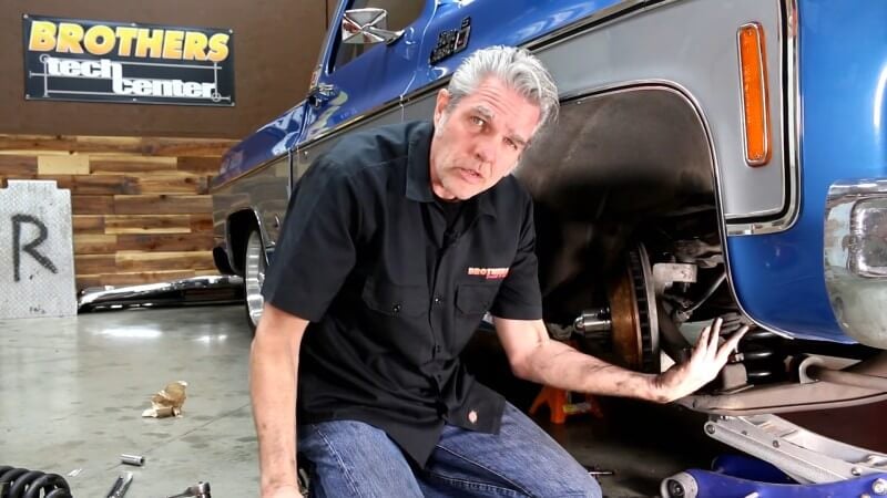 A mechanic kneeling next to a classic Chevy truck while pointing at the newly reconnecting tie rod end