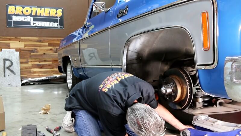 A mechanic kneeling next to a classic Chevy truck while centering a hydraulic jack underneath the lower control arm on the passenger side