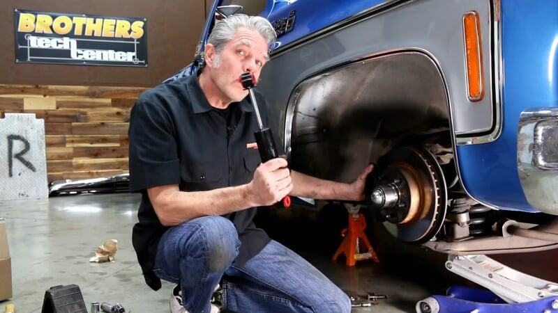 A mechanic kneeling next to a classic Chevy truck while holding the front shock absorber up to the camera