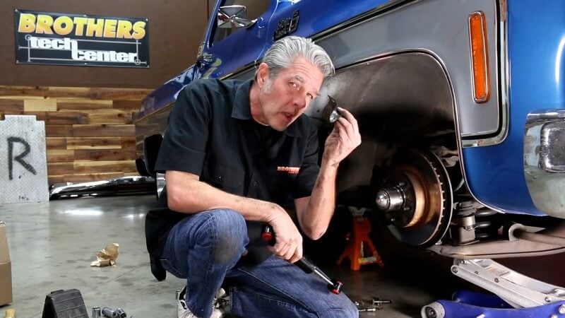 A mechanic kneeling next to a classic Chevy truck while holding the front sway bar bracket up to the camera