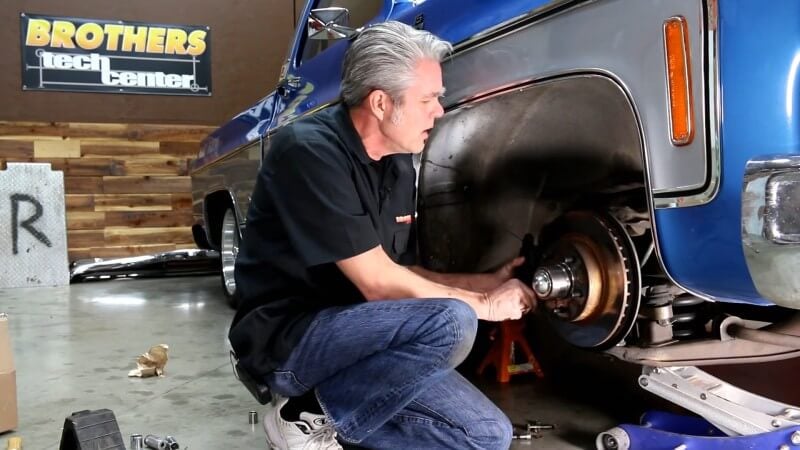 A mechanic kneeling next to a classic Chevy truck while sliding the front brake caliper off of the rotor