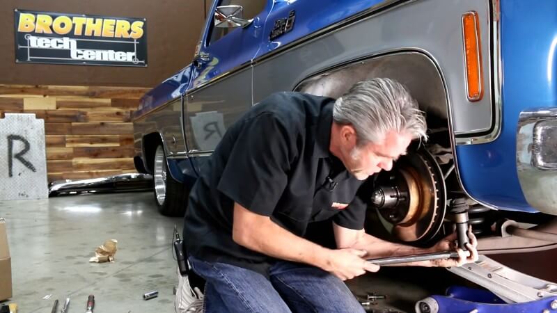 A mechanic kneeling next to a classic Chevy truck while using a breaker bar to loosen the tie rod end nut without fully removing it