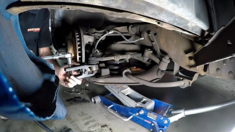 A mechanic kneeling next to a classic Chevy truck while using a tie rod end puller to and a ratchet to separate the tie rod end from the steering arm