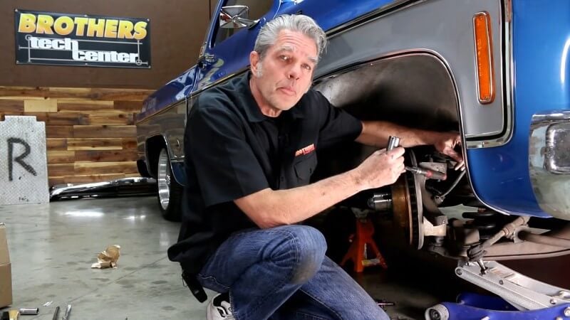 A mechanic kneeling next to a classic Chevy truck while using the end of a ratchet to point at the upper ball joint nut