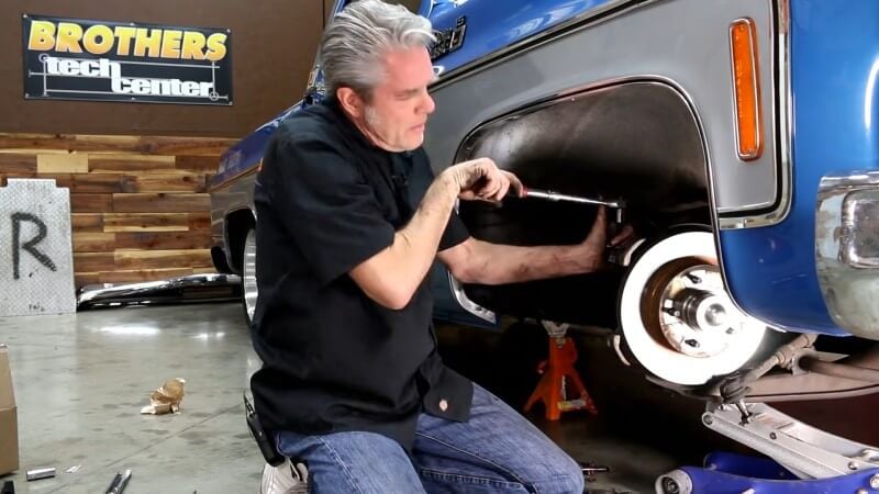 A mechanic kneeling next to a classic Chevy truck while using a breaker bar and a ball joint separator to release the upper ball joint from the spindle