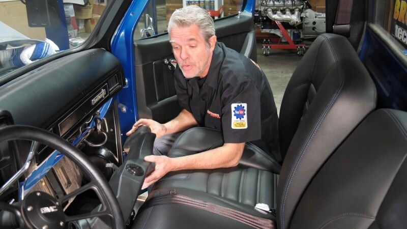 A mechanic sitting in the bed of the truck holding black plastic air conditioning components while looking at the camera