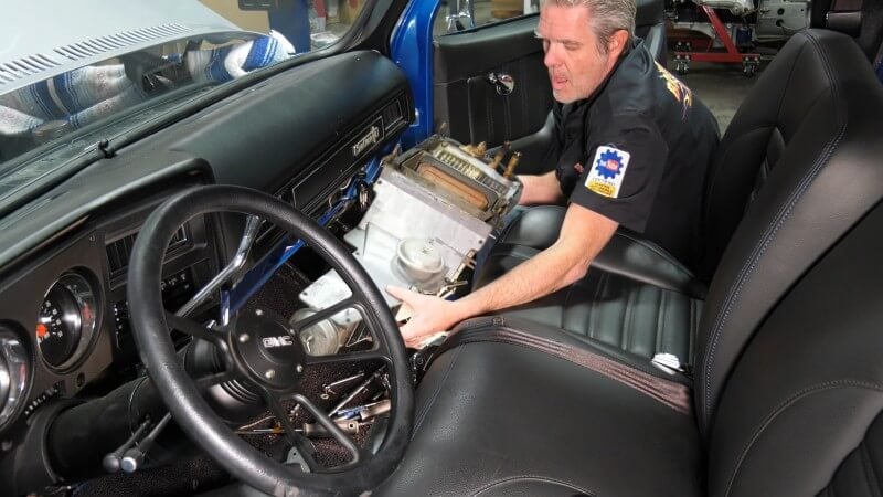 A mechanic sitting in the cab of a classic Chevy truck while pulling the heater box out from under the dashboard