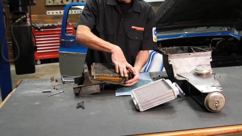 A mechanic standing in front of a table holding the heater core assembly uses his hands to remove the heater core brakets