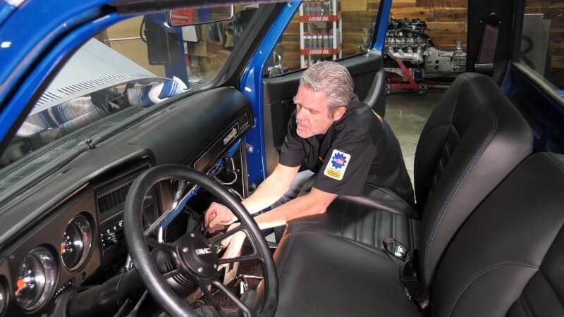 A mechanic sitting the cab of a classic Chevy truck and using his hands to reconnect the vacuum lines and cables underneath the dashboard in the passenger side footwell