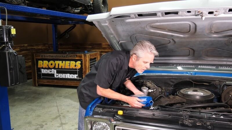 A mechanic leaning over the engine bay of a classic Chevy truck while pouring distilled water into the radiator