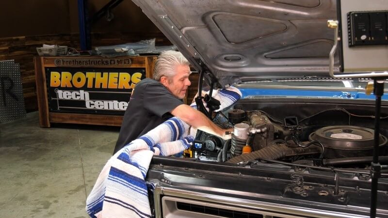 A wide angle shot of a mechanic leaning over the front fender of a classic Chevy truck and reaching into the engine bay to remove a heater hose clamp