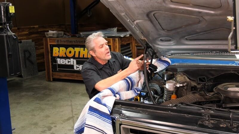 A wide angle shot of a mechanic leaning over the front fender of a classic Chevy truck and holding a disconnected heater hose up to the camera and pointing at it