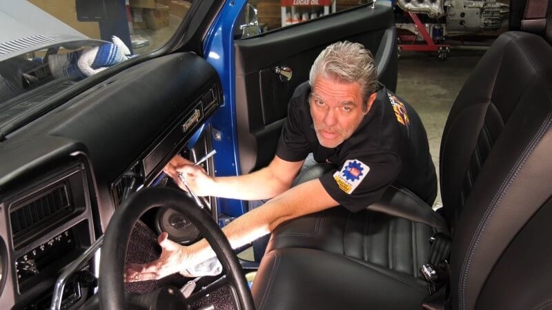 A mechanic sitting in the cab of a classic Chevy truck while gesturing at where the glove box was before removal