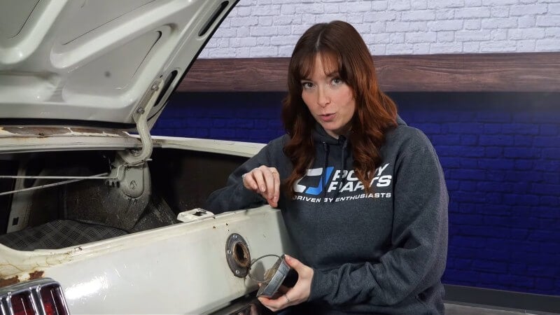 A female mechanic looking at the camera while removing the gas cap of a white classic Ford Mustang with her hands