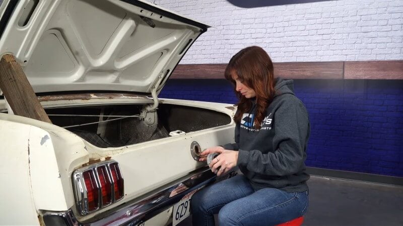 A female mechanic pulling the factory gas cap off of the trunk lid of a white classic Ford Mustang