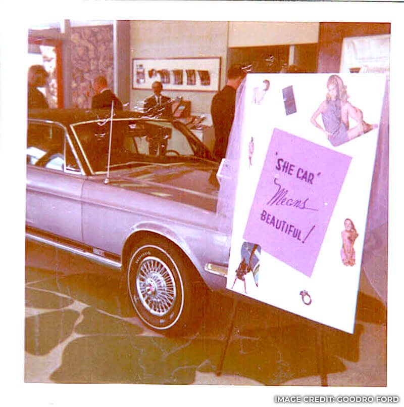 Vintage photo of the Evening Orchid Mustang beside sign that says “She Car Means Beautiful”