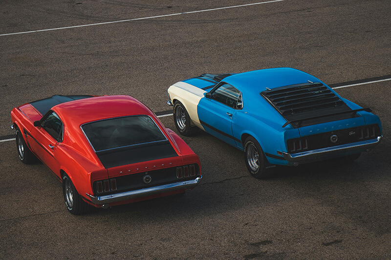 A red classic Mustang Mach 1 and a blue classic Mustang Mach 1 parked in a parking lot