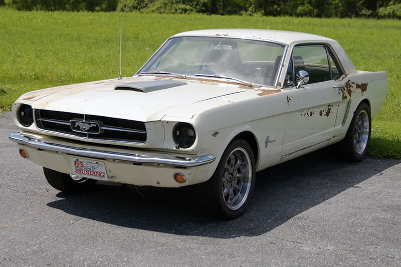 A white, slightly rusty classic Mustang parked in a parking lot in front of a field
