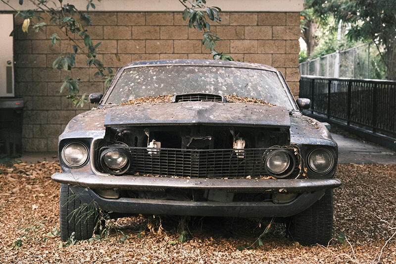 A rusty, dirty, and damaged classic Mustang parked under a tree in front of a garage